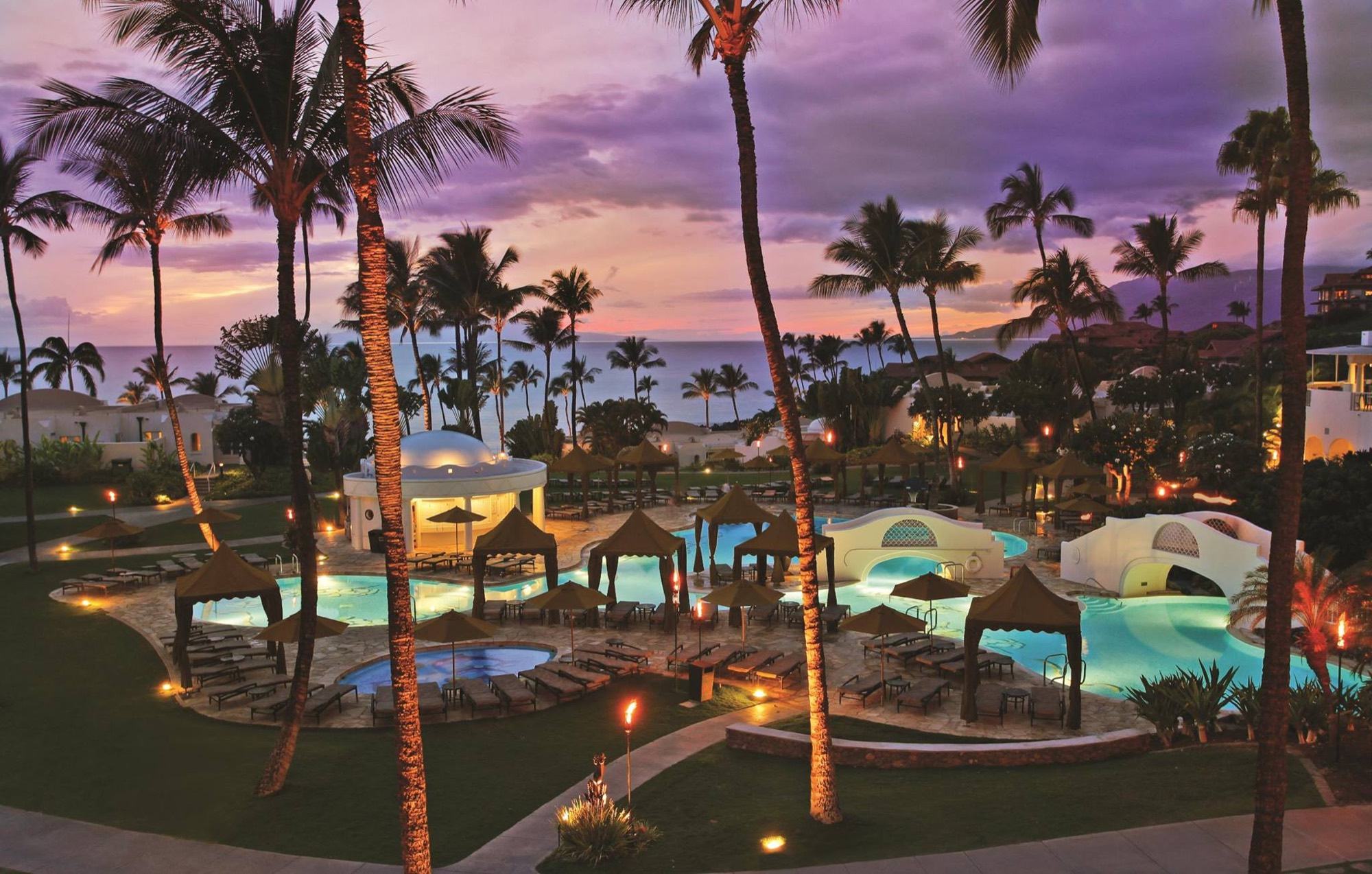 The lagoon pools at dusk beneath palms, with torches lit and the sky turning violet