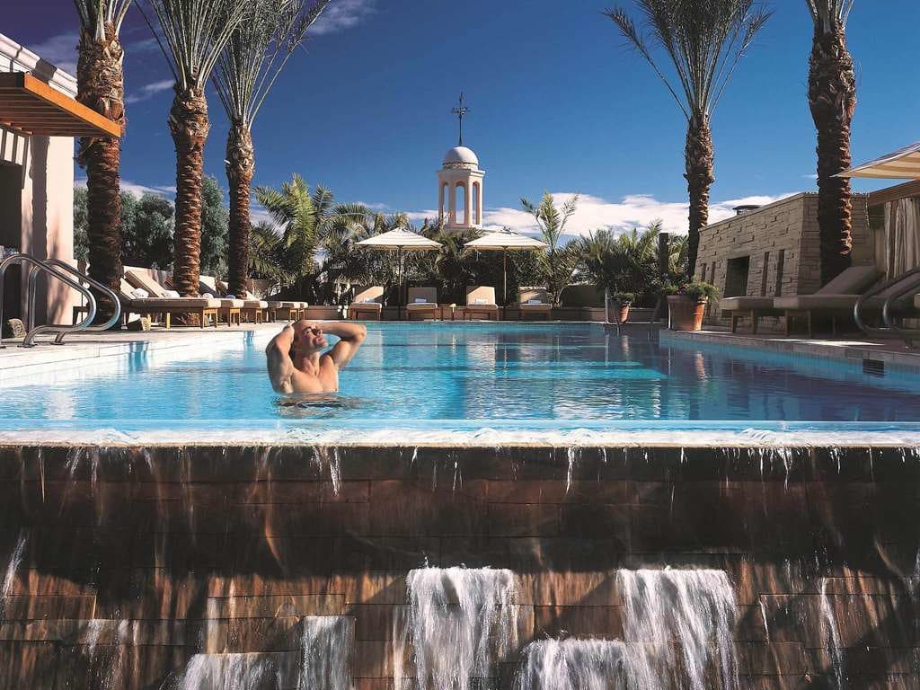 A swimmer at the edge of a pool that spills over a stone waterfall, with palms and a bell tower beyond