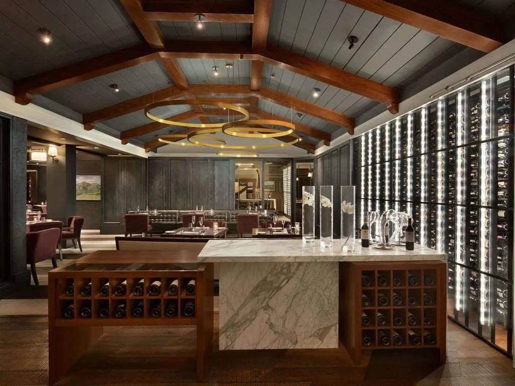A dining room beneath a beamed ceiling, beside a lit wall of wine bottles