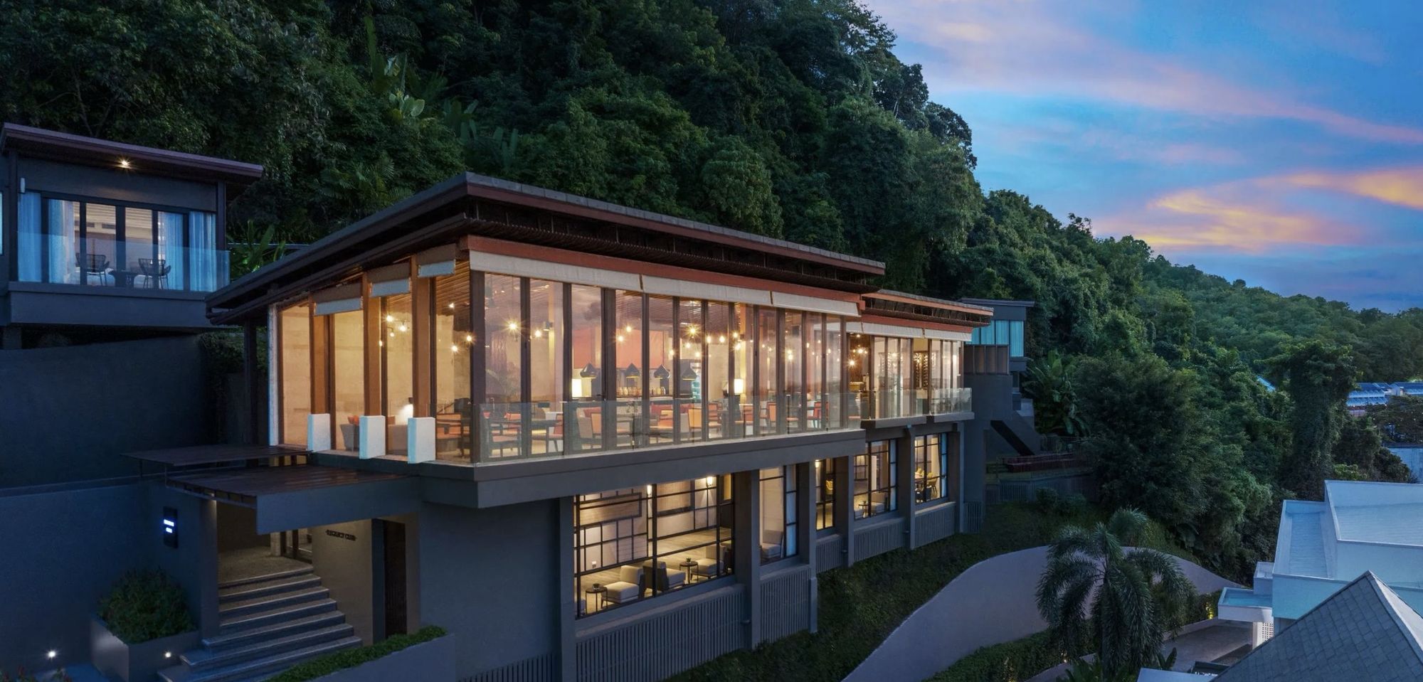 A glass-walled pavilion lit up on the green hillside at dusk