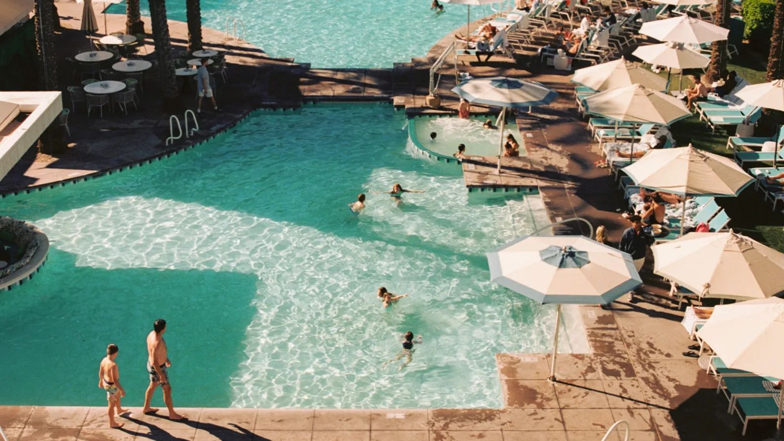 Swimmers and striped umbrellas around a hotel pool on a sunny day